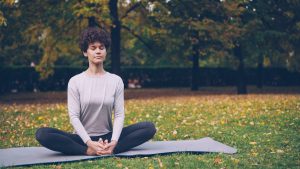 Flexible young lady is sitting on mat in yoga pose enjoying fresh air, peace and relaxation. Healthy lifestyle for urban people, active youth and autumn nature concept.